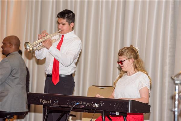 A student in a white button up shirt, a red tie and black pants plays the trumpet. To the right, another student in a white shirt and a red skirt plays the keyboard.